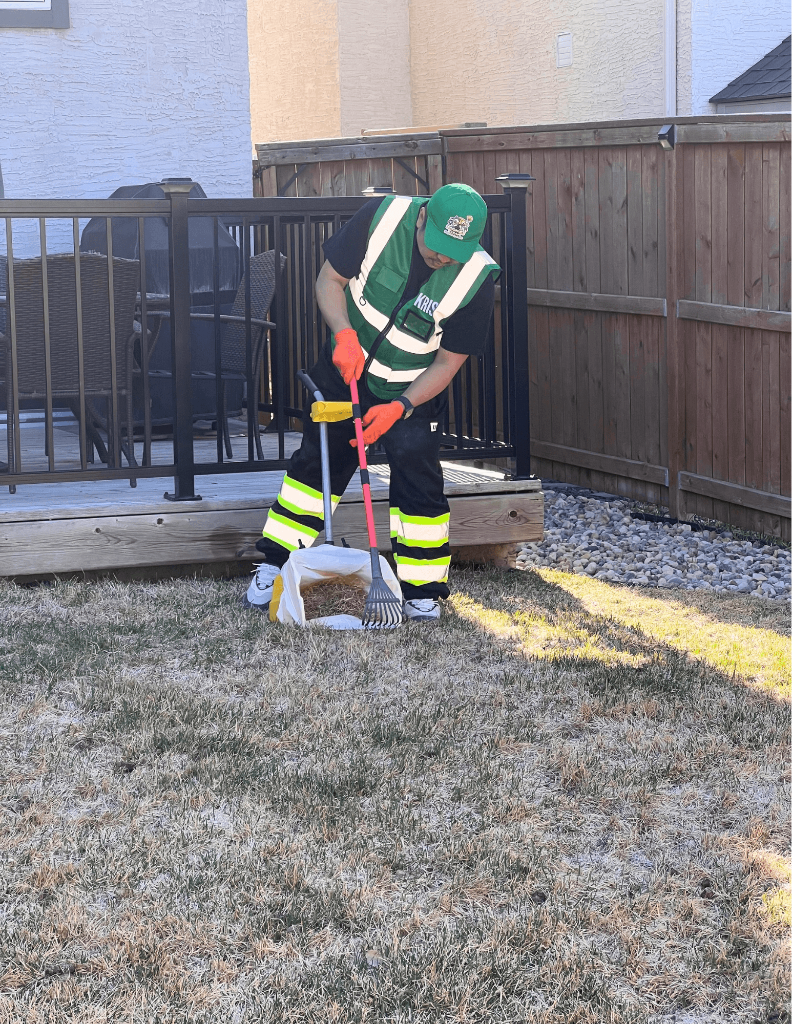 Worker in a green safety vest raking debris into a bag on a residential lawn.